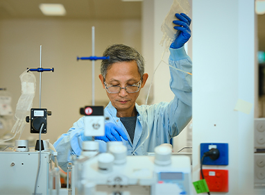 a scientist is working on some equipment and holding up a donation bag, they are wearing a blue gown and gloves