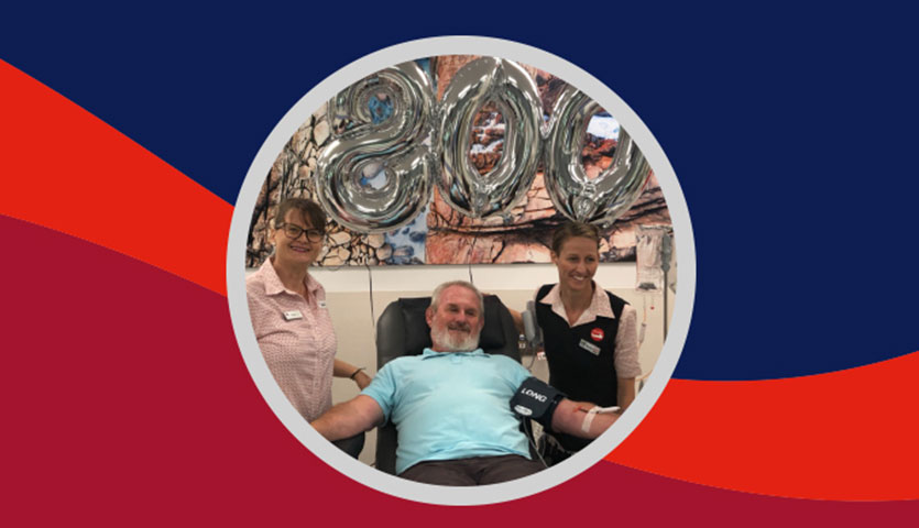 geoff is sitting in a chair and smiling during a donation, nurses stand either side of him and above them are silver coloured balloons in the number 800