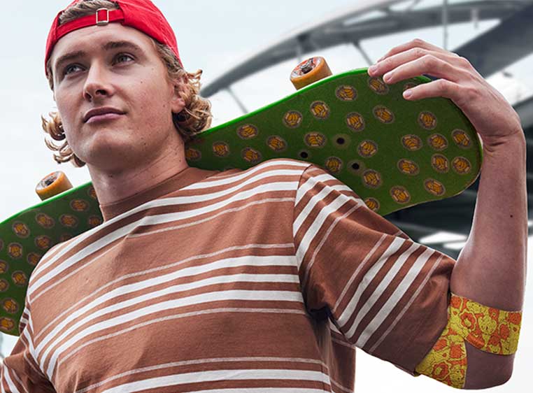 a young man is standing holding a skateboard over his shoulders, on his left elbow is a true colours designed bandage