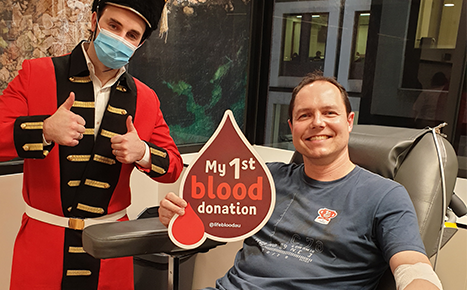 Man donating blood with a sign that reads my 1st blood donation next to a man wearing a cosutme of an english guard