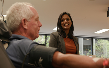 plasma donor Rowan is seated while giving a donation, recipient Dinushka is standing to his left and smiling