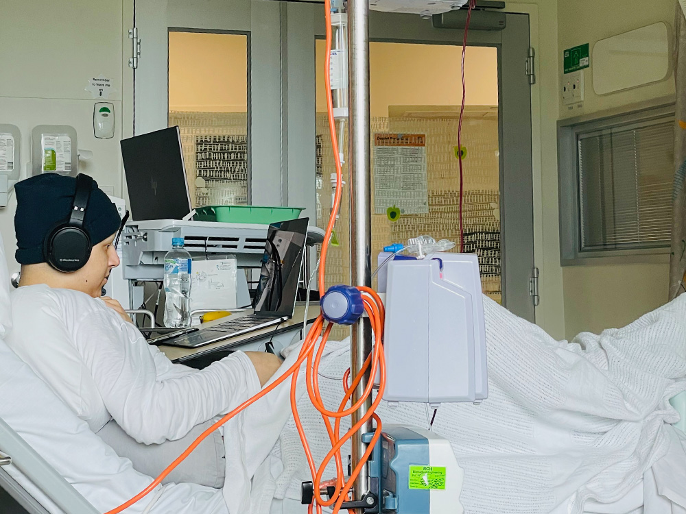 teenage boy wearing a beanie and headphones while looking at a laptop and receiving treatment for cancer