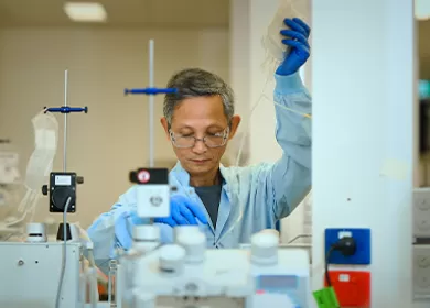 a scientist is working on some equipment and holding up a donation bag, they are wearing a blue gown and gloves