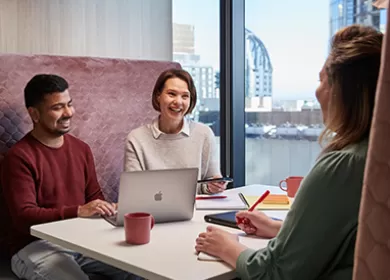 three work colleagues are sitting in a booth and smiling mid conversation, a laptop and notepads are on the table