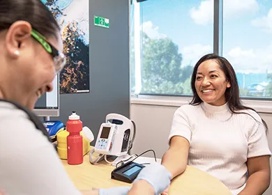 a woman is seated at a table smiling while a nurse performs a finger prick test