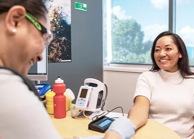 a woman is seated at a table smiling while a nurse performs a finger prick test