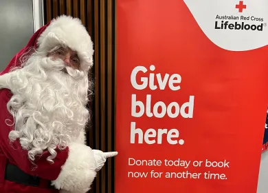 Man dressed as Santa in red suit and white beard pointing at sign which reads Give blood here 