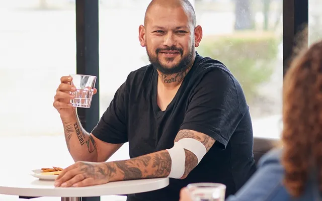 a man with tattoos seated at a table in a donor centre with a bandage around his elbow