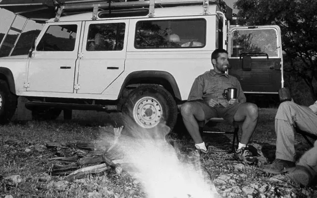 two men sitting on camp chairs around a fire chatting