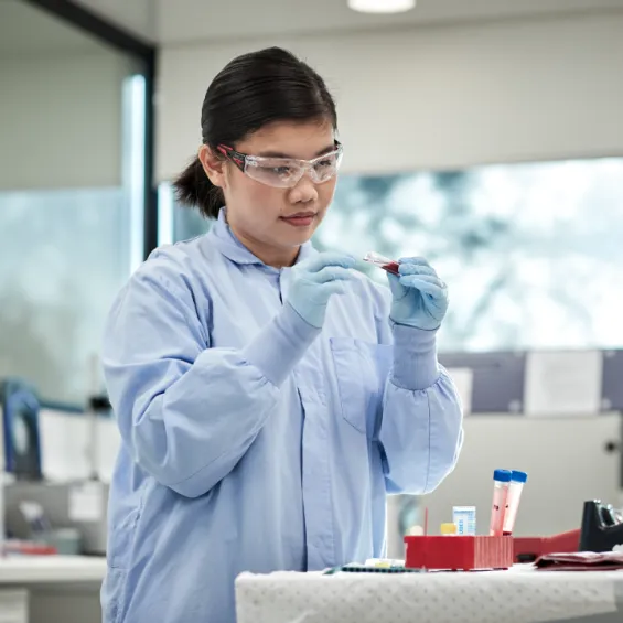 image of scientist in blue lab coat wearing safety glasses inspecting a test tube filled with blood