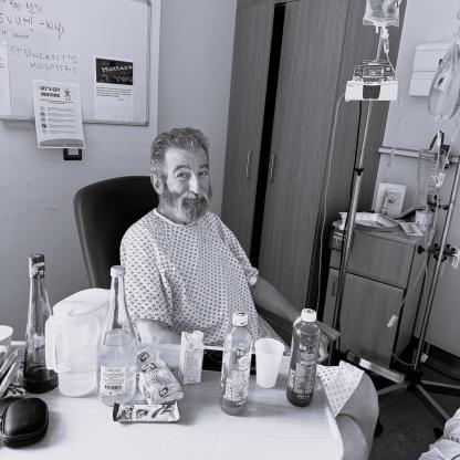 An older man with a grey beard sitting in a chair in a hospital room, he is wearing a hospital gown